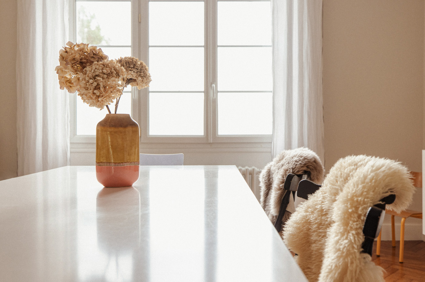 Maison Rambourg, vase en céramique rose et jaune contenant une fleur d'hortensia séchée posé sur la table du salon en plan large devant la fenetre